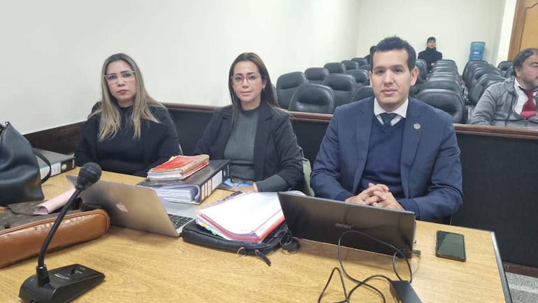 Tres personas en una mesa: mujer con blusa negra, mujer con chaqueta negra y top verde, hombre con saco azul y camisa blanca.