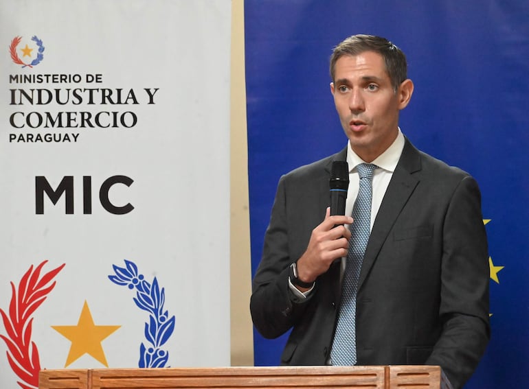 Marco Riquelme en traje oscuro y corbata, hablando con micrófono, frente a un atril del Ministerio de Industria y Comercio de Paraguay.