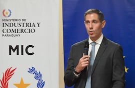 Marco Riquelme en traje oscuro y corbata, hablando con micrófono, frente a un atril del Ministerio de Industria y Comercio de Paraguay.