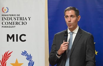 Marco Riquelme en traje oscuro y corbata, hablando con micrófono, frente a un atril del Ministerio de Industria y Comercio de Paraguay.