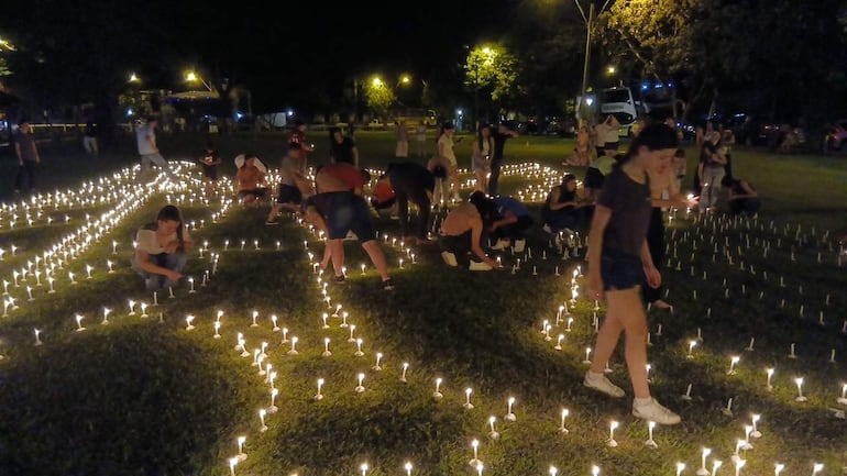 Múltiples personas en parque colocan velas encendidas sobre el césped, bajo iluminación de faroles, creando un ambiente conmemorativo.
