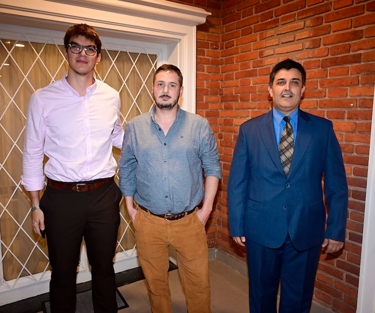 Tres hombres de pie en un salón con paredes de ladrillo; uno lleva traje azul, otro una camisa gris y el tercero una camisa rosa.