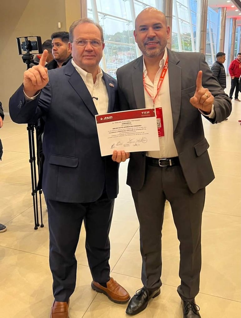 Camilo Pérez, con abrigo negro, levanta un certificado con expresión feliz, junto a Óscar Rodríguez en un evento formal.