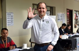 Hombre sonriente con camisa blanca y dedo marcado por tinta, levantando la mano en un local de votación organizado.