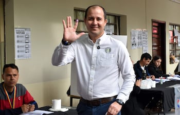 Hombre sonriente con camisa blanca y dedo marcado por tinta, levantando la mano en un local de votación organizado.