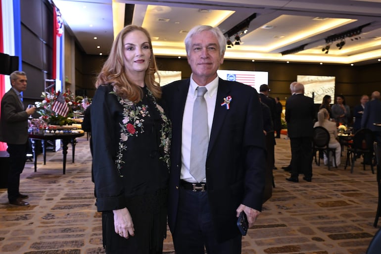 Mujer con blusa negra bordada y hombre con saco oscuro, posando en un salón decorado con flores y mesas elegantes.