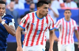 Diego Gómez con camiseta blanca y rayas rojas, corriendo en el campo, rodeado de un ambiente de estadio y un balón de fútbol.