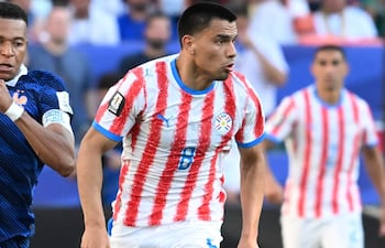Diego Gómez con camiseta blanca y rayas rojas, corriendo en el campo, rodeado de un ambiente de estadio y un balón de fútbol.