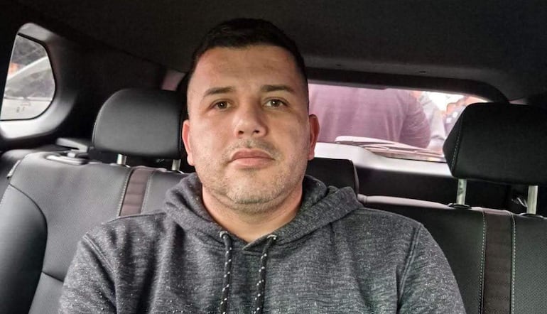 Hombre con sudadera y cabello corto sentado en vehículo, expresando tranquilidad, con siluetas difusas detrás.