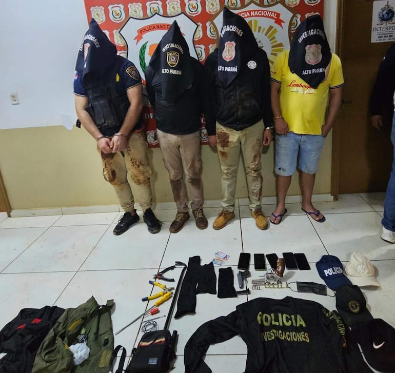 Cuatro personas con capuchas y ropa oscura, esposadas en una oficina policial, rodeadas de objetos incautados.