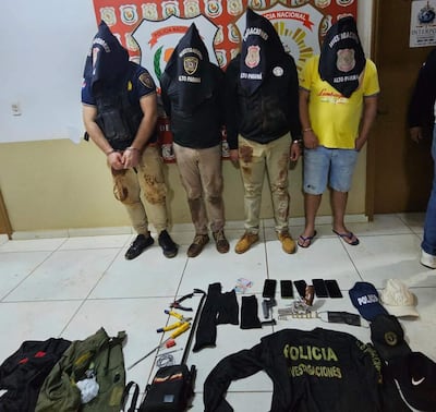 Cuatro personas con capuchas y ropa oscura, esposadas en una oficina policial, rodeadas de objetos incautados.