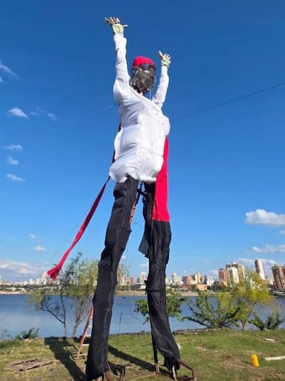 Muñeco gigante vestido de blanco y negro con gorro rojo, brazos levantados, frente a una ciudad con edificios altos.