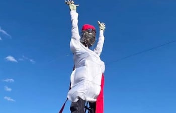 Muñeco gigante vestido de blanco y negro con gorro rojo, brazos levantados, frente a una ciudad con edificios altos.
