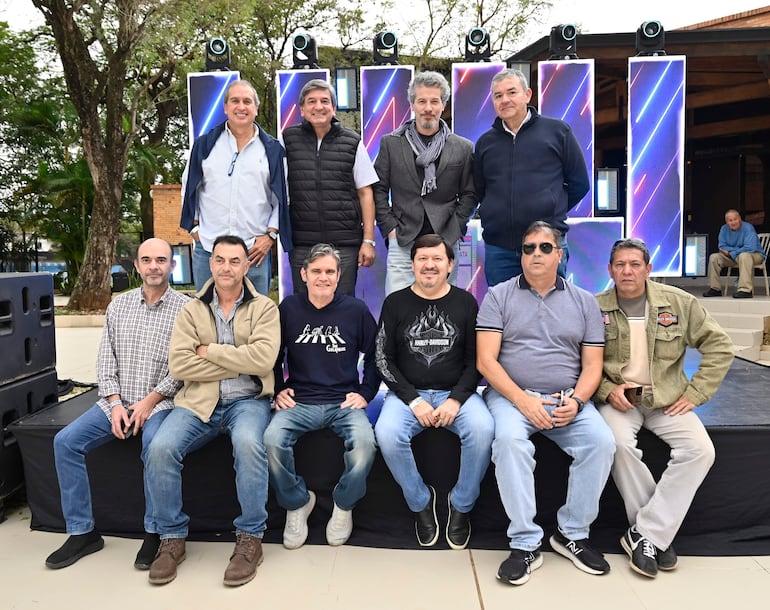 Grupo de diez hombres posando sonrientes, algunos sentados en una tarima negra, con luces de escenario de fondo.