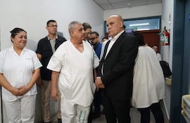 Isaías Fretes en bata blanca y Bachi Núñez en traje oscuro conversan, mientras una enfermera sonríe al fondo en un hospital.