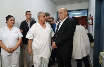 Isaías Fretes en bata blanca y Bachi Núñez en traje oscuro conversan, mientras una enfermera sonríe al fondo en un hospital.