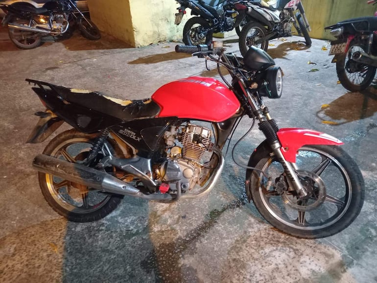Motocicleta negra y roja estacionada en un entorno húmedo, sin personas visibles y rodeada de otras motos.