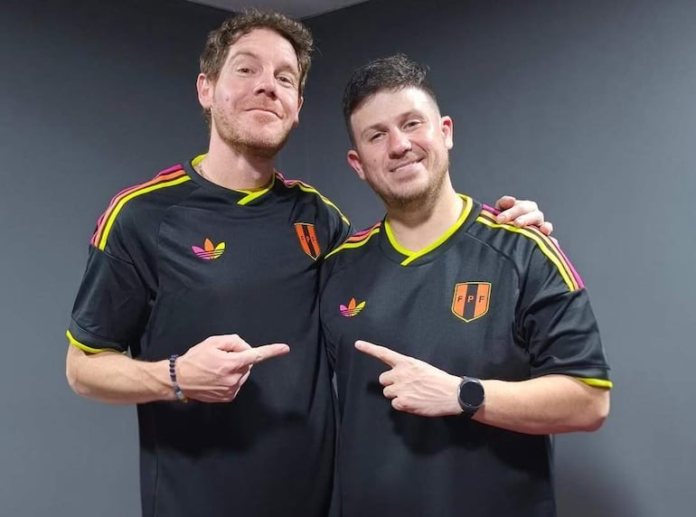 Diego Vicos y Brian Caruso sonríen en camisetas negras con detalles amarillos y lilas, apuntando hacia el centro de la habitación gris.