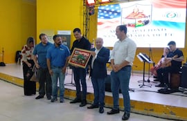Grupo de personas en salón amarillo, un hombre en traje sostiene un cuadro decorativo, mientras otros observan con expresión feliz.