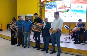 Grupo de personas en salón amarillo, un hombre en traje sostiene un cuadro decorativo, mientras otros observan con expresión feliz.