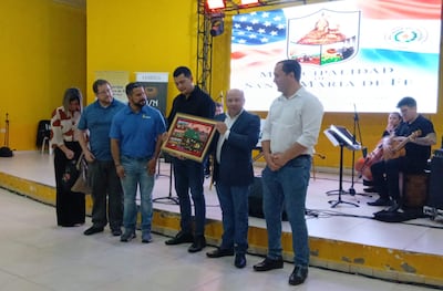 Grupo de personas en salón amarillo, un hombre en traje sostiene un cuadro decorativo, mientras otros observan con expresión feliz.