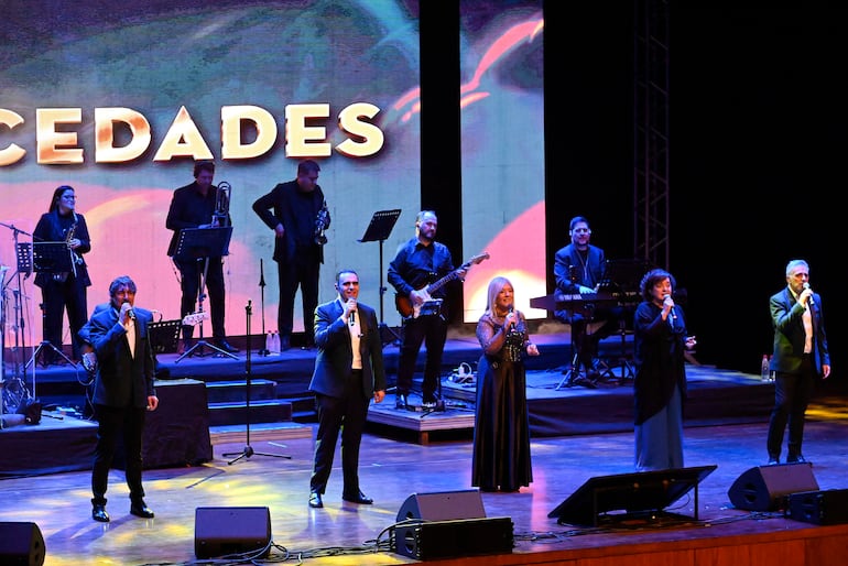 Cinco cantantes de Mocedades en trajes formales, con dos hombres cantando y tres mujeres participando, en un escenario con luces.