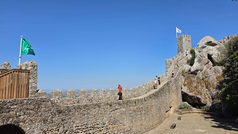 Turistas en camiseta roja y dos hombres en la muralla del castillo, disfrutando de un día soleado y del paisaje.