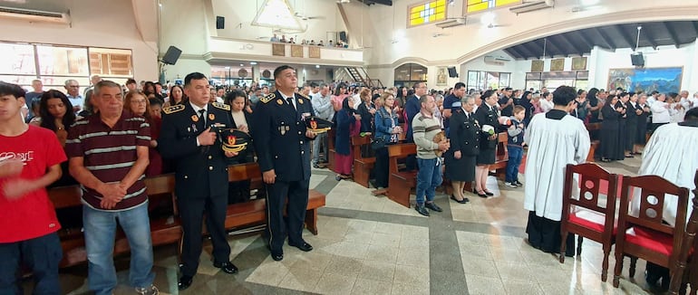 Varias personas en actitud de oración, con hombres uniformados al frente sosteniendo un objeto y bancos de madera en el interior del lugar.