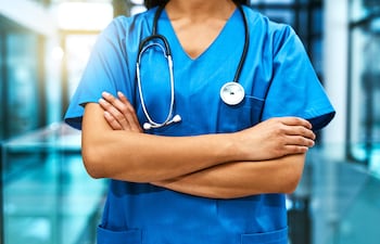 Médica en uniforme azul, con brazos cruzados y estetoscopio, mostrando postura confiada en ambiente clínico.