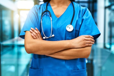 Médica en uniforme azul, con brazos cruzados y estetoscopio, mostrando postura confiada en ambiente clínico.