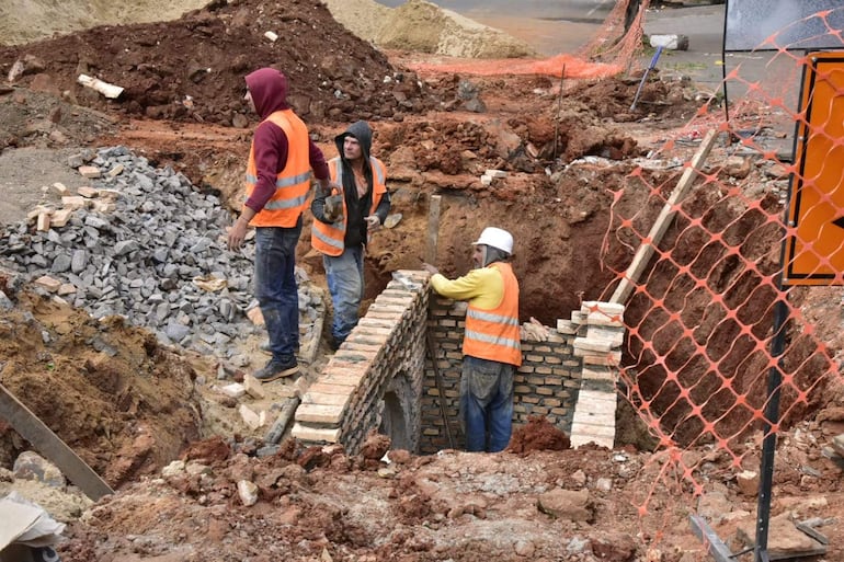 Cuatro hombres en construcción, uno excava con ladrillos, otro lleva casco blanco y chalecos naranjas en un terreno de tierra y piedras.