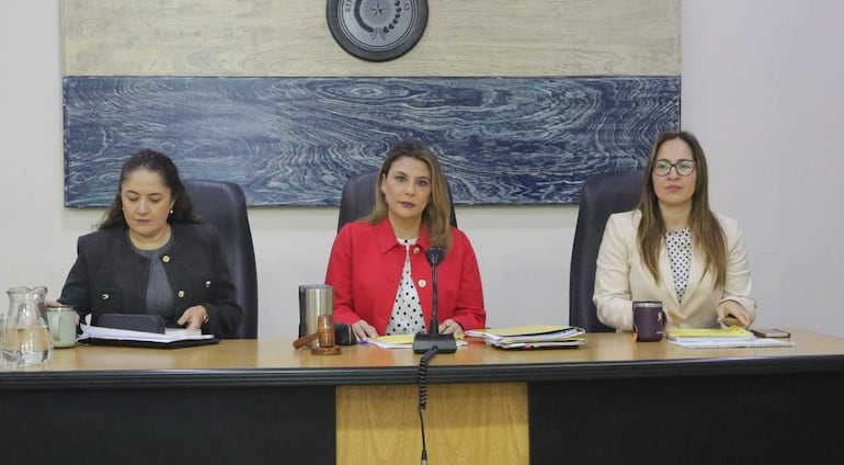 Tres mujeres sentadas en mesa, una en chaqueta roja habla, otra en blanco mira a la cámara, y la de negro se concentra en un documento.