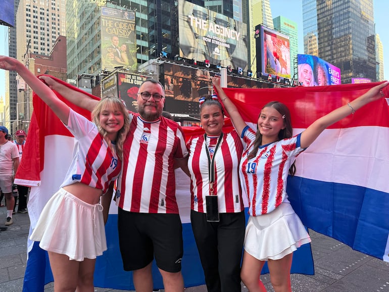 Cuatro jóvenes en camiseta de rayas rojas y blancas sostienen una bandera paraguaya con expresiones de alegría en un ambiente festivo.