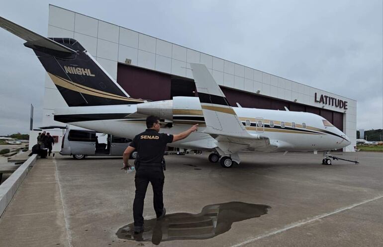 Hombre con camiseta negra 'SENAD' señala un avión privado. Aeropuerto Silvio Pettirossi con edificio 'LATITUDE' en el fondo.