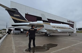 Hombre con camiseta negra 'SENAD' señala un avión privado. Aeropuerto Silvio Pettirossi con edificio 'LATITUDE' en el fondo.