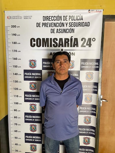 Hombre serio, cabello corto, con camiseta azul y jeans, frente al cartel de la Comisaría 24° con fondo amarillo.