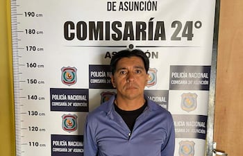 Hombre serio, cabello corto, con camiseta azul y jeans, frente al cartel de la Comisaría 24° con fondo amarillo.