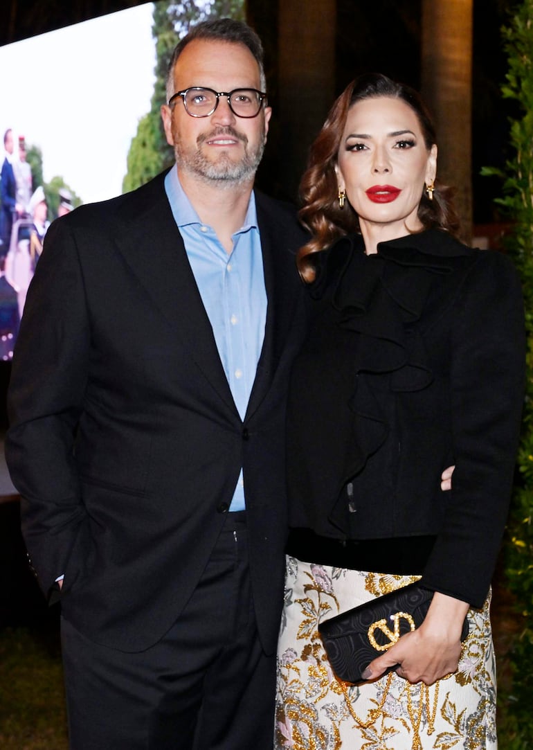Mujer con atuendo negro y labios rojos sonríe junto a hombre con saco negro y camisa azul en ambiente iluminado.