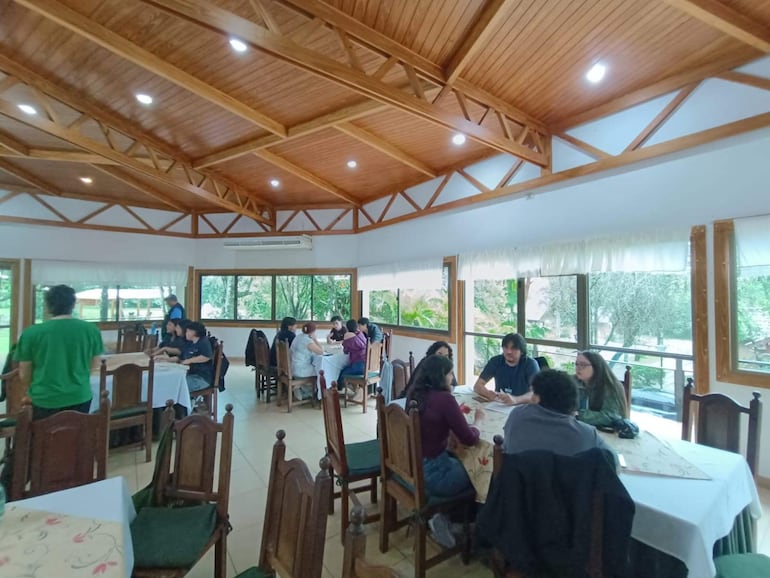 Grupo de jóvenes conversando animadamente en un restaurante, con una persona sirviendo a la izquierda. Ambiente iluminado y alegre.