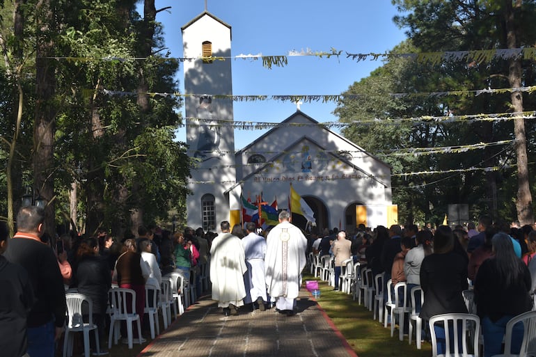 Tres sacerdotes de espaldas en misa al aire libre, rodeados de 100 asistentes, en un ambiente decorado con banderas en un día soleado.