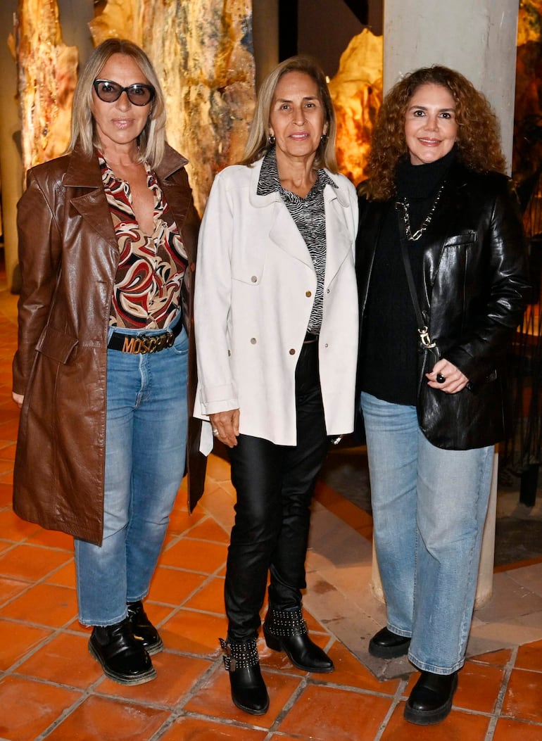 Tres mujeres sonrientes: una con abrigo de cuero marrón, otra con chaqueta blanca y otra con abrigo negro, en ambiente moderno.