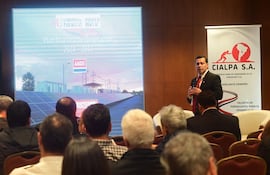 Hombre de pie con traje oscuro y corbata roja presenta información, mientras el público lo observa atentamente en un salón de conferencias.