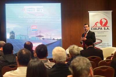 Hombre de pie con traje oscuro y corbata roja presenta información, mientras el público lo observa atentamente en un salón de conferencias.