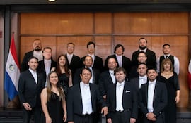 Grupo de 18 personas vestidas formalmente posan en un salón, con banderas de Paraguay detrás y un director al frente con batuta.