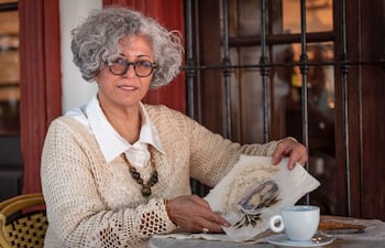 Mujer con cabello rizado y canoso, en suéter claro, sostiene papel decorado y observa atentamente en ambiente acogedor.