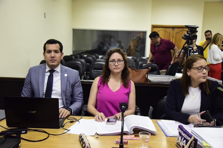 Tres personas sentadas en mesa de reunión: hombre en traje gris, mujer en blusa rosa con teléfono, otra mujer en blusa blanca concentrada.