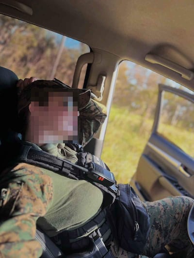 Soldado con rostro parcialmente cubierto, vestido de camuflaje y chaleco táctico, descansando en un vehículo en un paisaje natural.