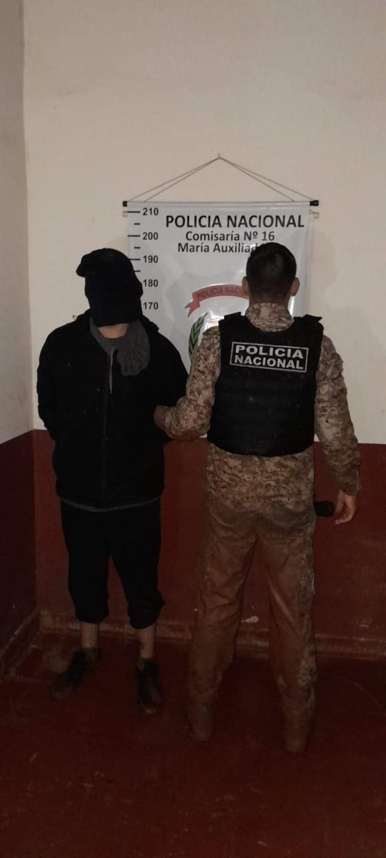 Hombre con chaqueta negra de espaldas, custodiado por policía en chaleco táctico en comisaría.