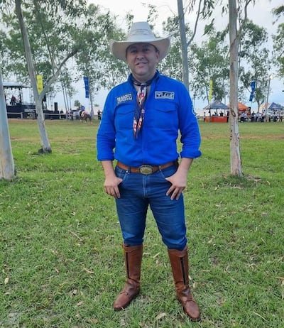 Lider Amarilla, sonriente, con camisa azul y sombrero blanco, vestido de jinete en un evento rodeado de árboles y césped.