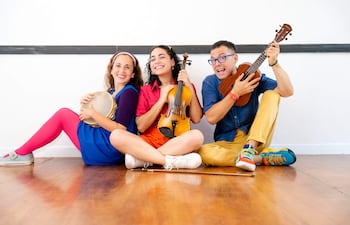 Mujeres con vestido azul y rizado sosteniendo un violín y un pandero, mientras hombre con camisa azul toca ukelele, todos sonrientes.